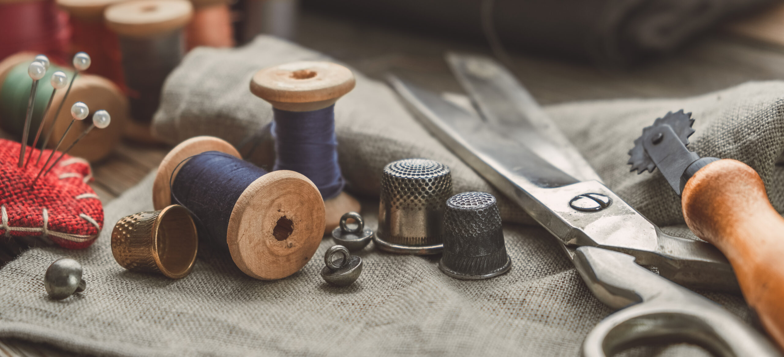 Retro sewing items: tailoring scissors, cutting knife, thimble, wooden thread spools, cushion for including pins, fabrics and sewing accessories.